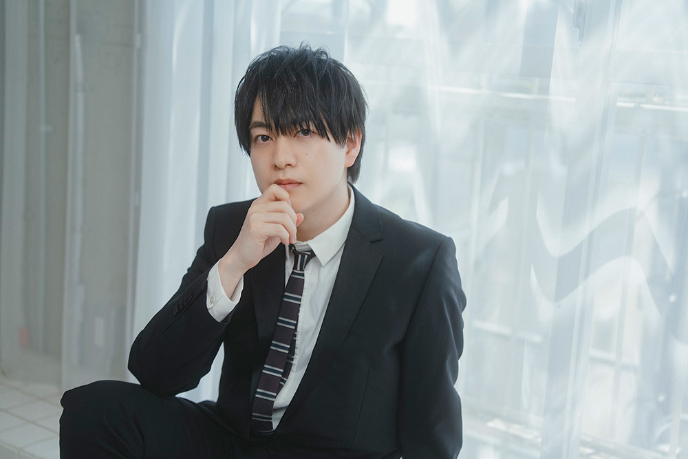 スーツ姿で窓際に腰掛ける小林さん。