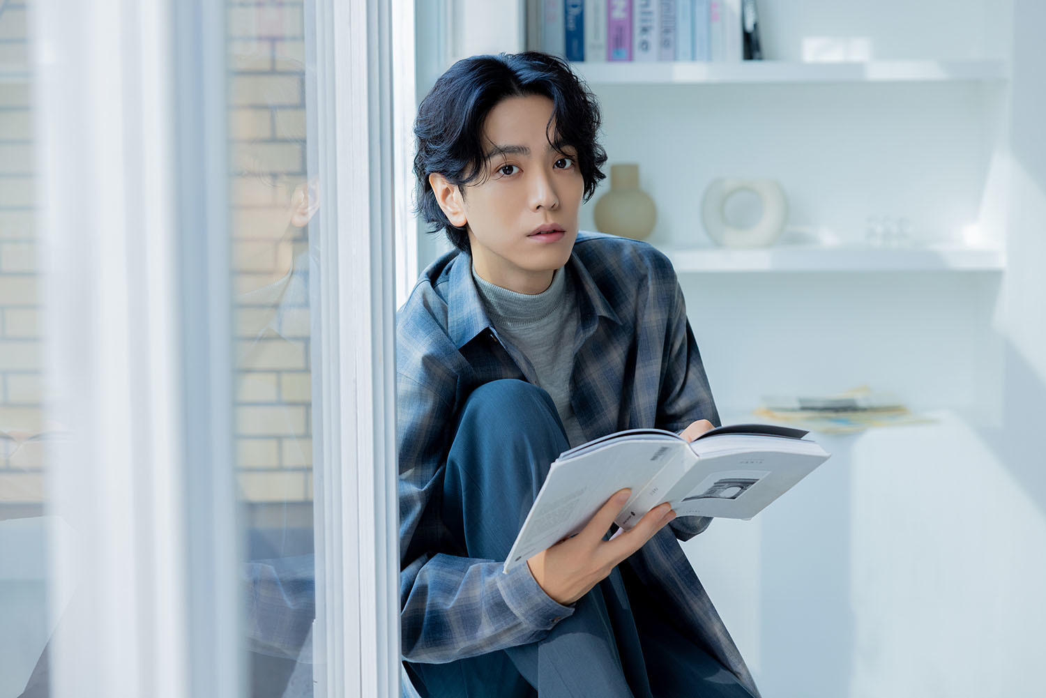 窓辺で本を手にしカメラを見る黒羽さん。